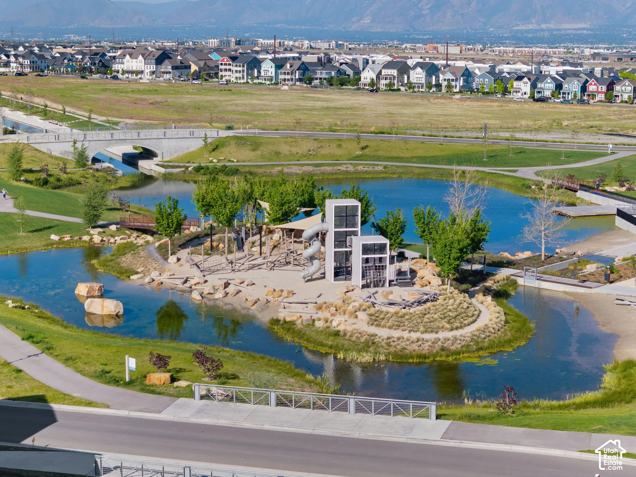 Expansive community park featuring a large pond with a central play area, pedestrian pathways, and a backdrop of residential architecture