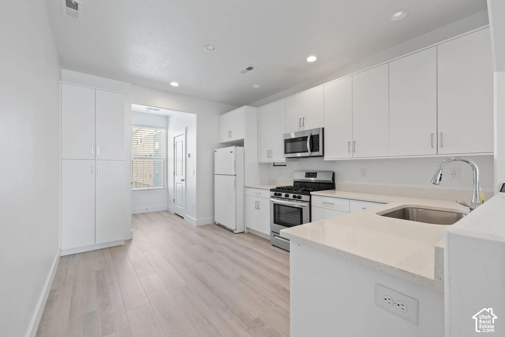Modern kitchen featuring white cabinetry, light-colored quartz countertops, stainless steel appliances, a stainless steel sink with a gooseneck faucet, and wood-finish flooring, abundant cabinet space and natural light