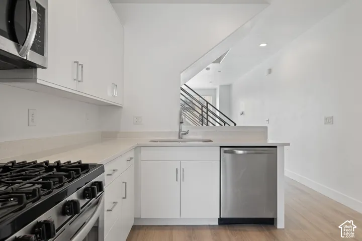 Modern kitchen featuring white cabinetry, light-colored quartz countertops, stainless steel appliances, a stainless steel sink with a gooseneck faucet, and wood-finish flooring, abundant cabinet space and natural light