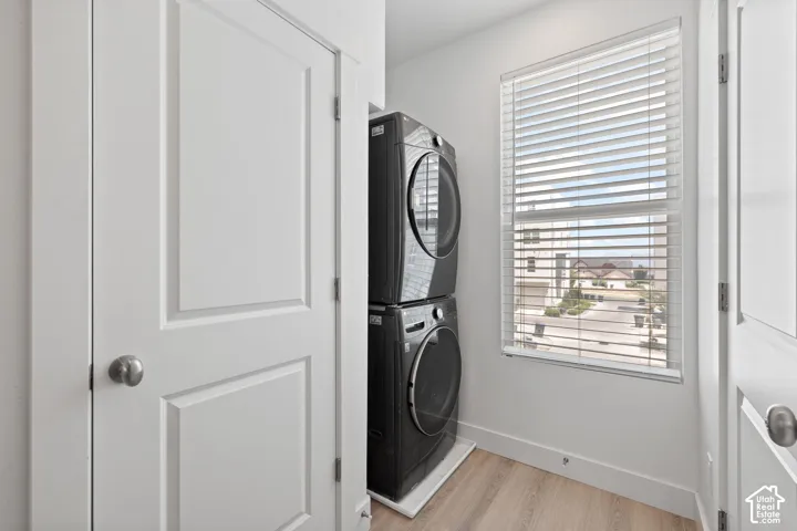 Dedicated laundry area featuring stacked appliances, a large window with white blinds, wood-finish flooring, and white paneled doors