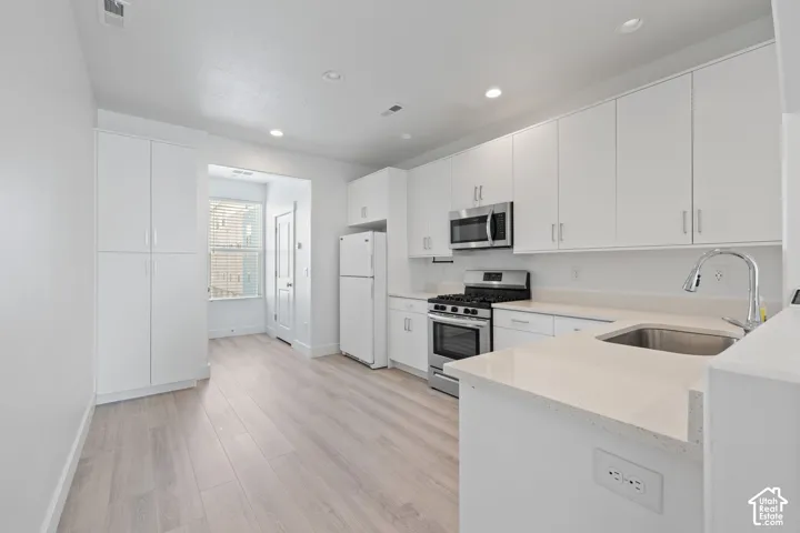 Modern kitchen featuring white cabinetry, light-colored quartz countertops, stainless steel appliances, a stainless steel sink with a gooseneck faucet, and wood-finish flooring, abundant cabinet space and natural light