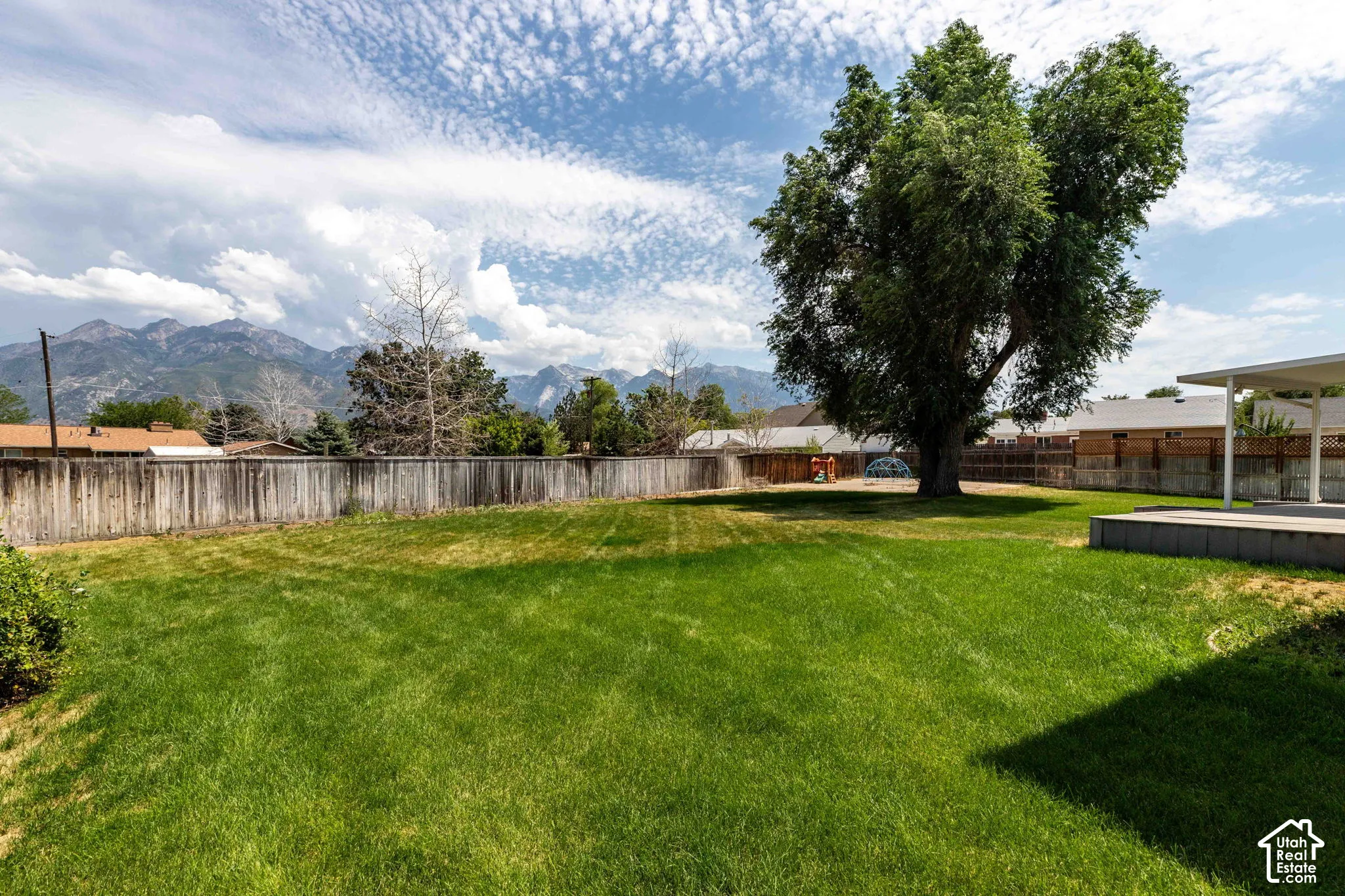 Expansive fenced backyard featuring a large mature tree, green lawn, a patio with an overhead cover, and mountain scenery