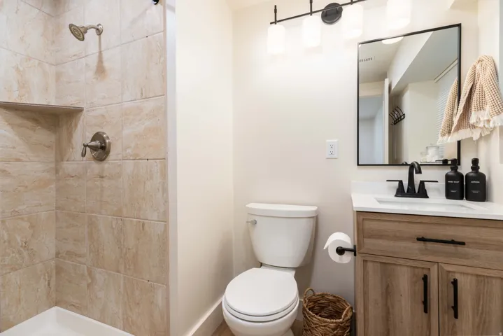 Bathroom featuring a walk-in shower with tiled walls and a built-in shelf, a toilet, and a vanity with wood-finish cabinetry, a white countertop, and a black-framed mirror