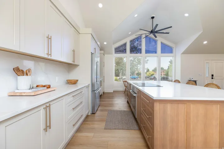 Vaulted ceiling kitchen featuring a large central island with integrated cooktop, light wood-finish flooring, full-height white cabinetry with gold-tone hardware, and a window wall with expansive exterior views