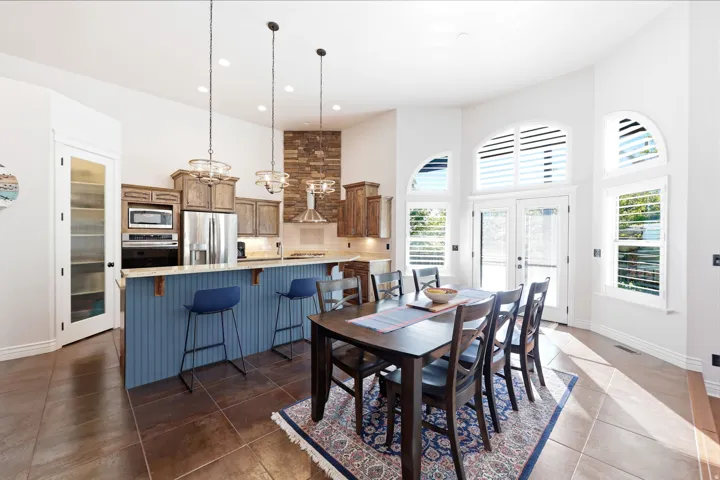 Spacious kitchen and dining area featuring vaulted ceilings, stainless steel appliances, a breakfast bar island with beadboard paneling, tiled flooring, and multiple arched windows with plantation shutters