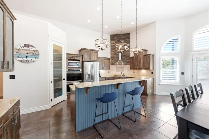 Spacious kitchen featuring a large central island with beadboard paneling, integrated stainless steel appliances, stacked stone range hood surround, wood cabinetry, and light-toned countertops