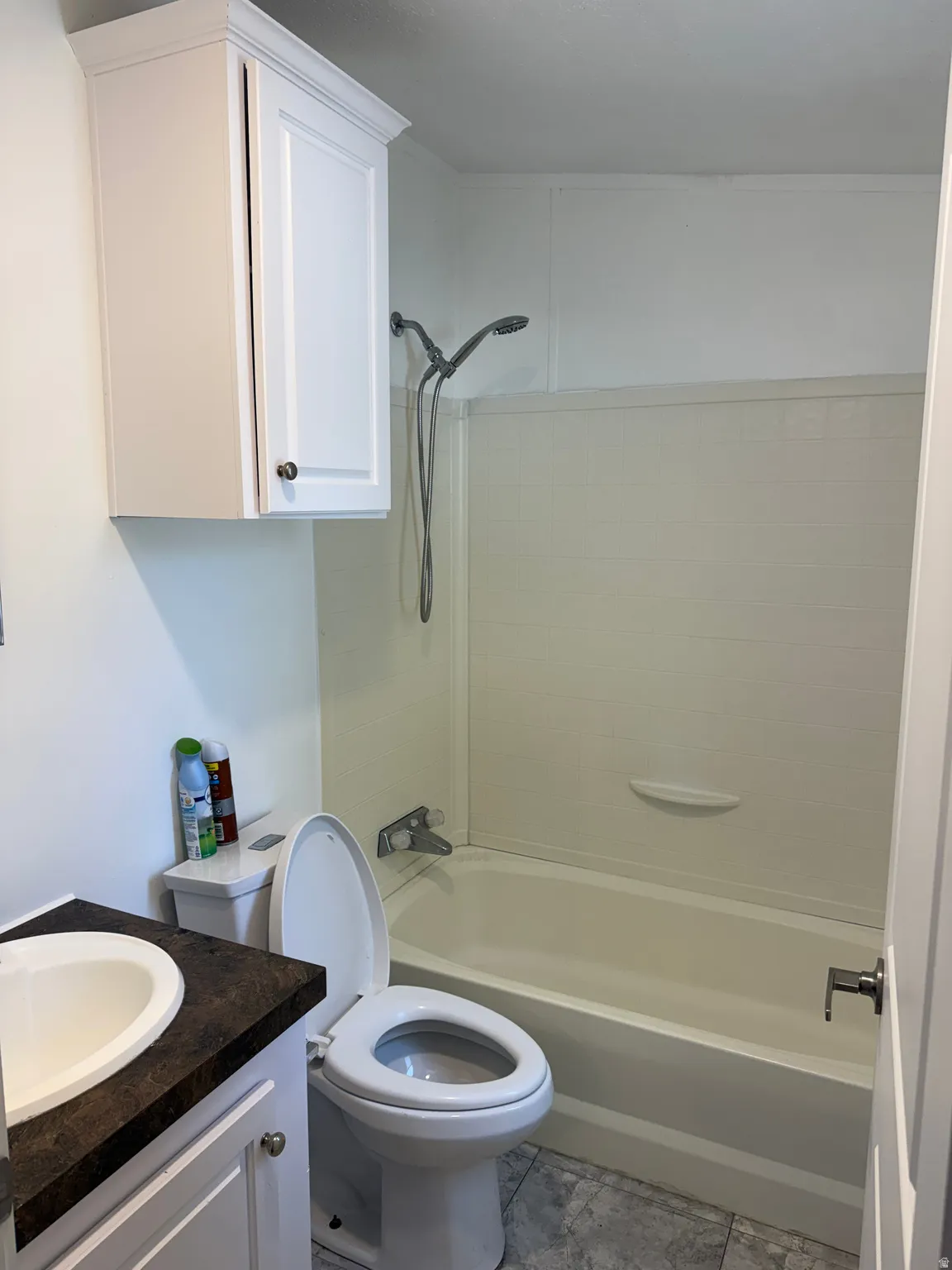 Compact bathroom featuring a white porcelain toilet, a white bathtub with an integrated shower, a white vanity with a dark countertop and an oval sink, and a white wall-mounted cabinet