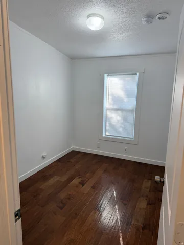 Compact room featuring rich wood-finish flooring, white baseboards, and a single window with blinds