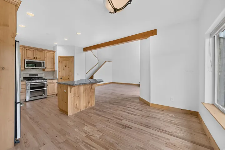 Open-concept living space featuring wood-finish flooring, a kitchen island with stone countertop, stainless steel appliances, recessed lighting, and an exposed wooden beam