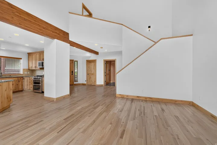 Open-concept interior featuring light wood-finish flooring, exposed wood ceiling beams, a kitchen with wood cabinetry and a stainless steel oven, and a partial staircase with wood trim