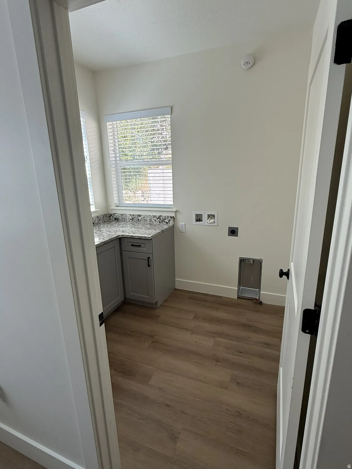 Functional utility space featuring a corner countertop with gray cabinetry, a window with blinds, and wood-finish flooring