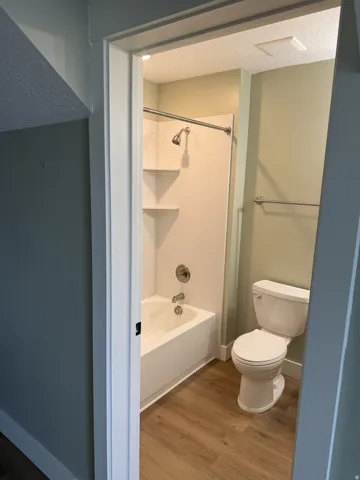 Bathroom featuring a white tub and shower combination with an integrated shelving unit, brushed nickel fixtures, and wood-finish flooring
