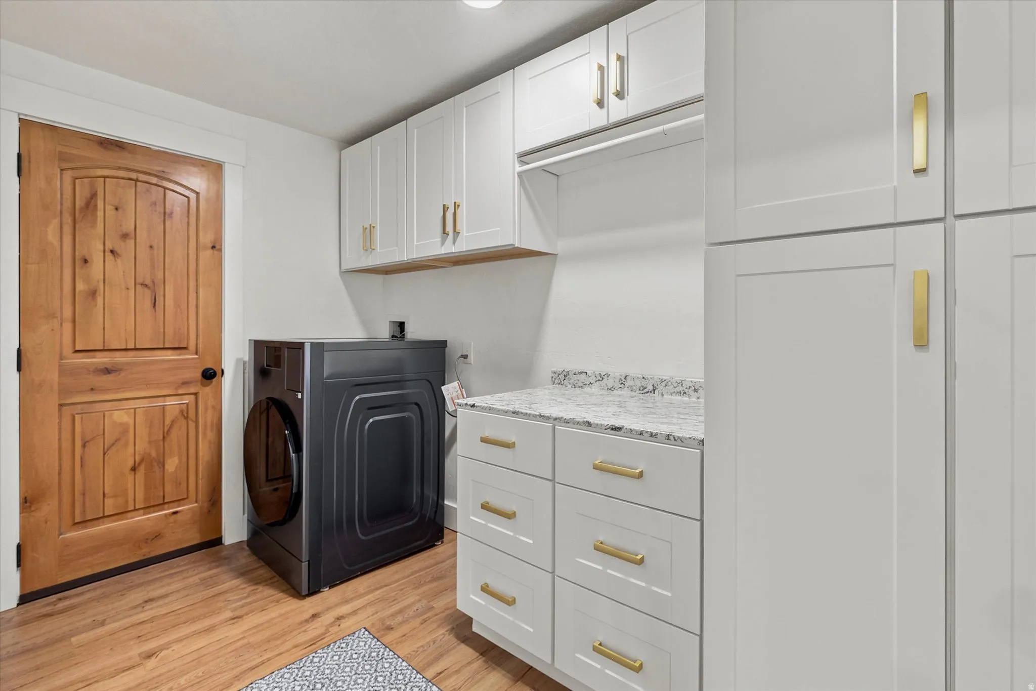 Laundry area featuring wood-finish flooring, a wood-panel door, white cabinetry with gold-finish hardware, and a light-toned countertop