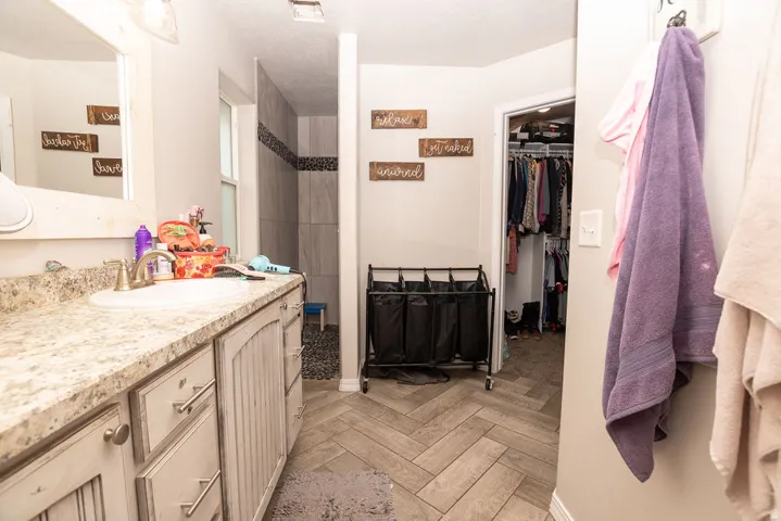 Bathroom featuring a single vanity with a countertop and undermount sink, a framed mirror, a tiled shower with a decorative accent band, and wood-finish flooring laid in a herringbone pattern