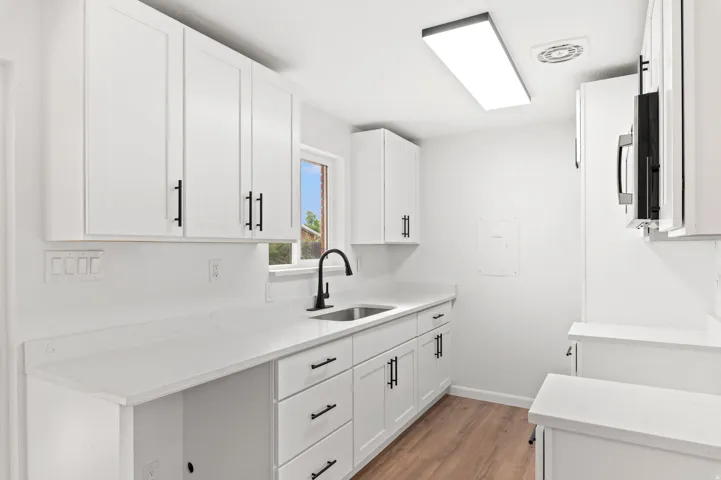 Bright kitchen featuring white shaker cabinetry with black hardware, white countertops, wood-finish flooring, a stainless steel sink with a matte black faucet, and a rectangular ceiling light fixture