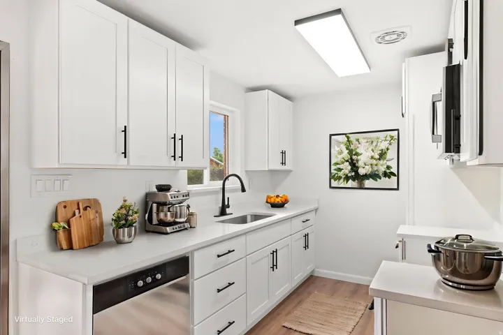 Galley kitchen featuring white cabinetry, light countertops, wood-finish flooring, a stainless steel dishwasher, and a contemporary black faucet w/ virtual staging