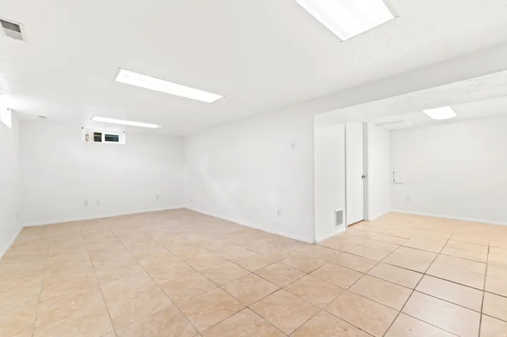 Spacious room featuring neutral tile flooring, white walls, multiple overhead fluorescent lighting fixtures, and a white interior door