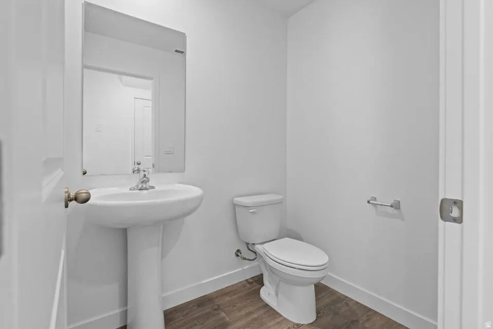 Pedestal sink with chrome faucet, wall-mounted mirror, toilet, wood-finish flooring, and white baseboards