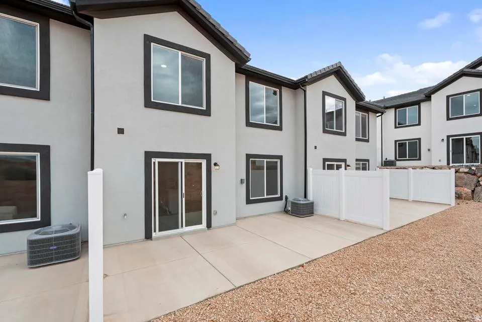 Rear exterior of a stucco townhouse featuring multiple windows with dark trim, a sliding glass door, and a concrete patio area