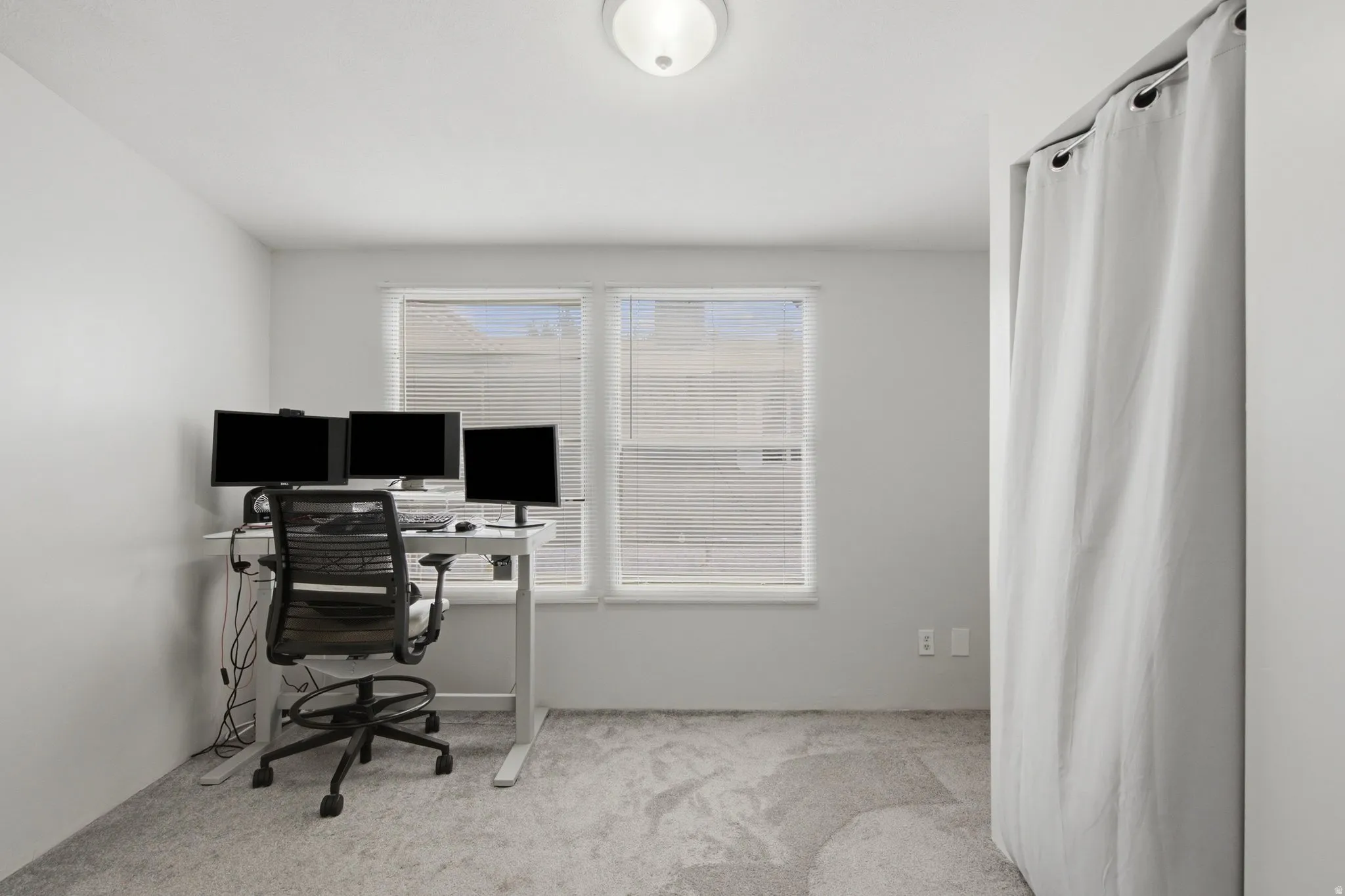 Room featuring light gray carpeting, two windows with blinds, a ceiling-mounted light fixture, and a curtained alcove