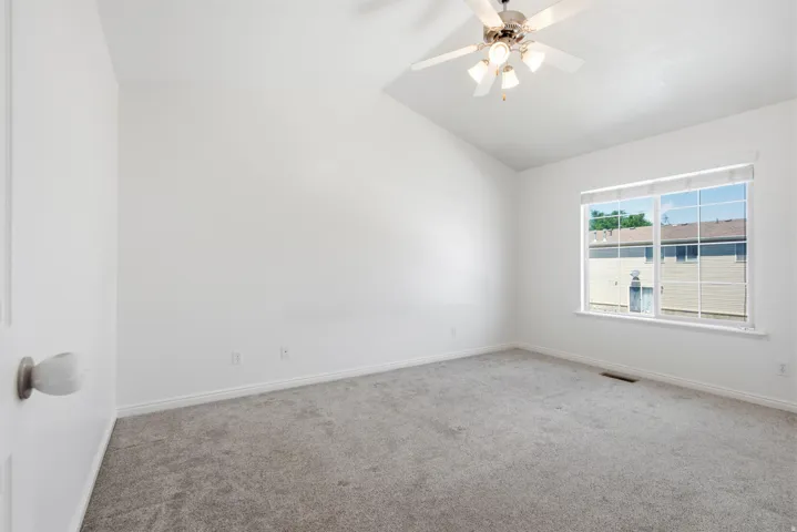 Bright room featuring vaulted ceilings, neutral gray carpeting, a large window, and a ceiling fan with integrated lighting