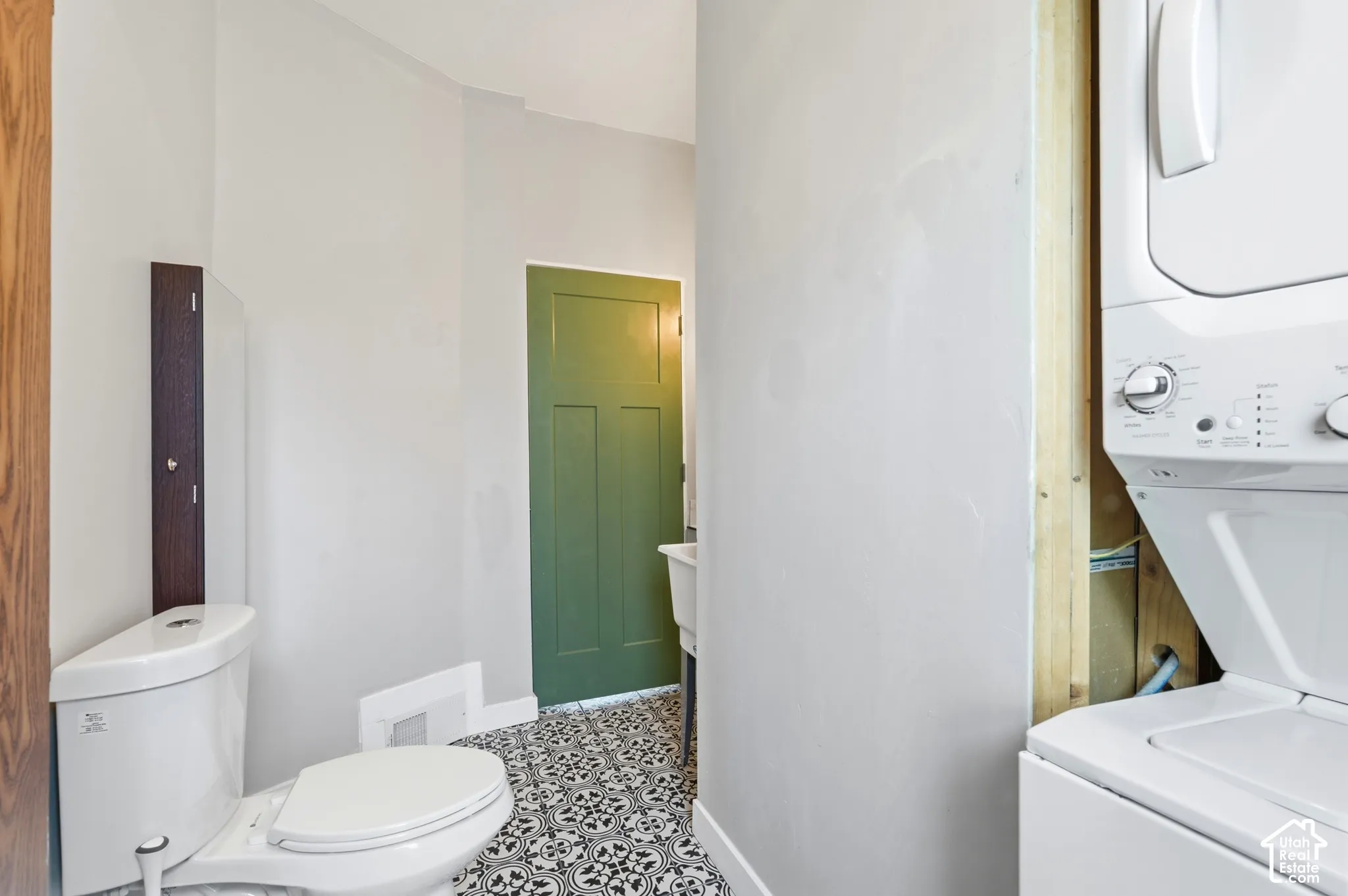 Floor-to-ceiling storage cabinet, white toilet, patterned floor tiles, green paneled door, and stacked laundry appliances