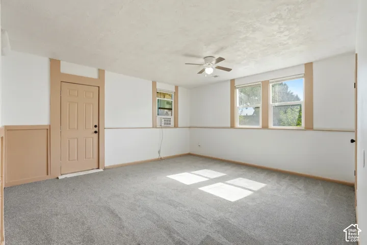 Spacious room featuring neutral carpeting, a paneled door, three windows with natural light, a ceiling fan with integrated lighting, and a wall-mounted air conditioning unit