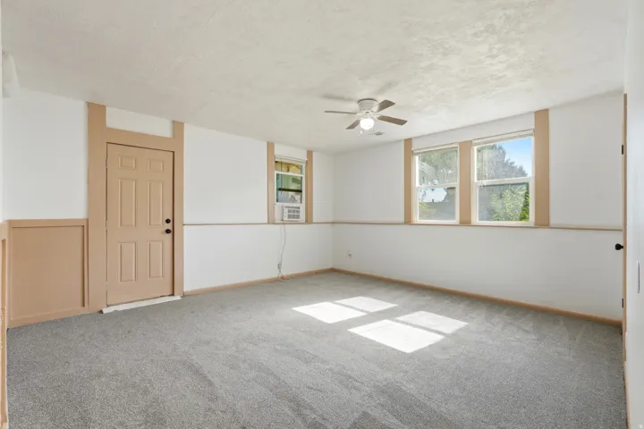 Spacious room featuring neutral carpeting, a paneled door, three windows with natural light, a ceiling fan with integrated lighting, and a wall-mounted air conditioning unit