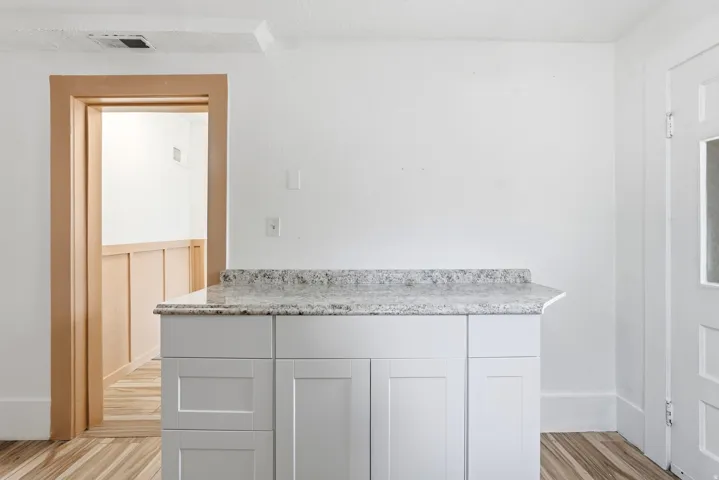 White cabinetry with a speckled countertop, light wood-finish flooring, white walls, and wood trim around an entryway