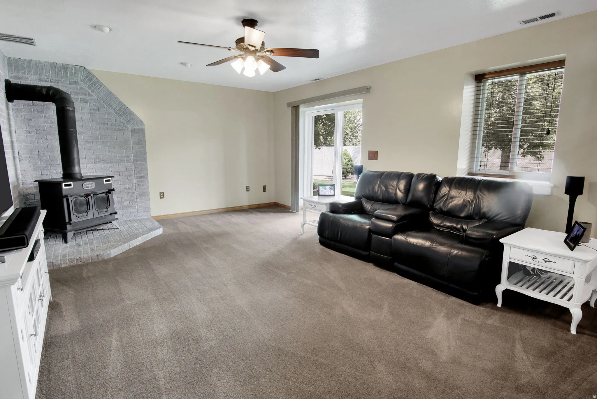 Spacious carpeted living area featuring a brick-clad wood-burning stove, a ceiling fan, a large window with blinds, and a sliding glass door providing exterior access