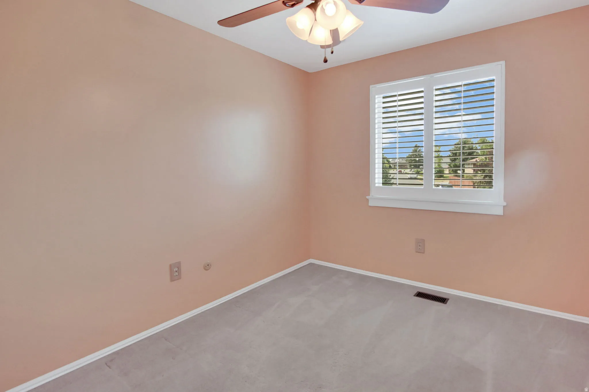 Room with soft pink walls, a white-framed window with plantation shutters, light gray carpeting, white baseboards, and a ceiling fan with integrated lighting