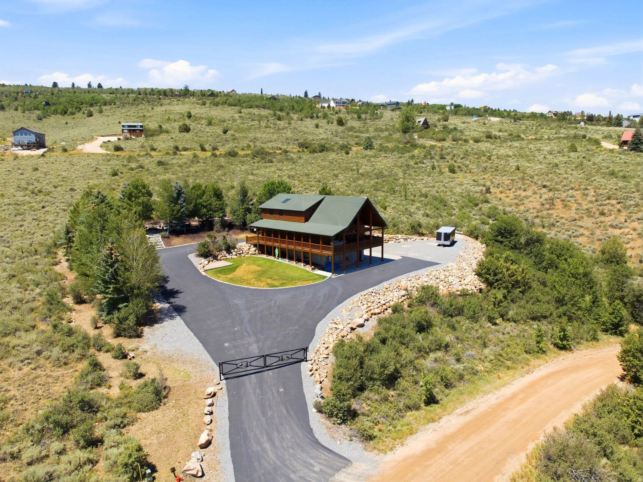 Rustic log home with a green roof, elevated deck, paved driveway, gated entrance, and surrounding natural landscaping