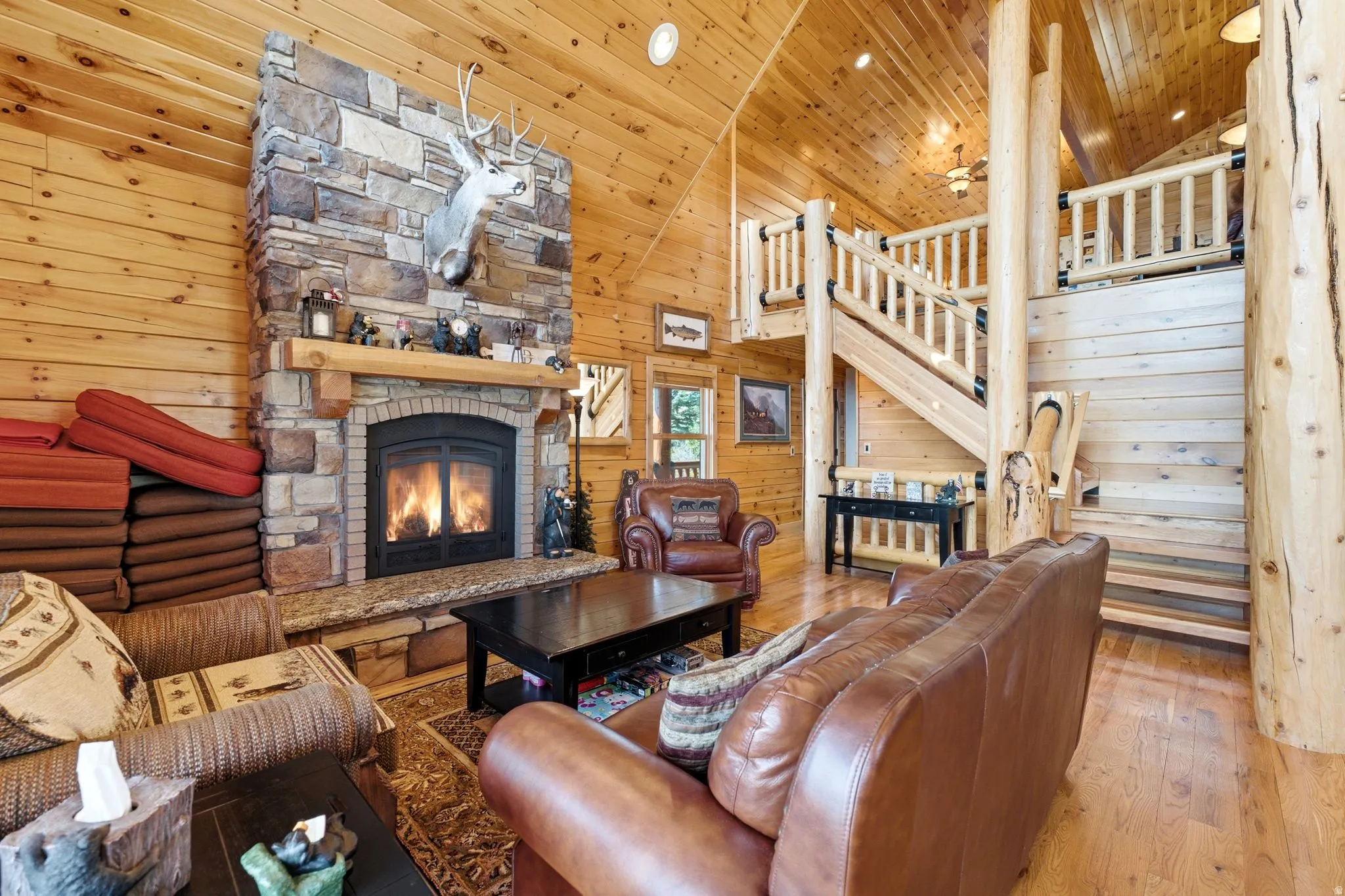 Vaulted ceiling living area featuring a stone fireplace with wood mantle, wood-finish flooring, and a log staircase leading to an upper level