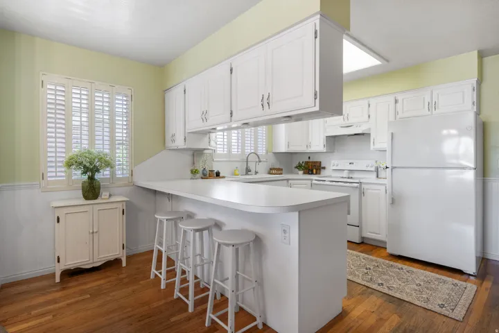 Kitchen featuring white cabinetry, a peninsula with seating, white appliances, wood-finish flooring, and a window with plantation shutters