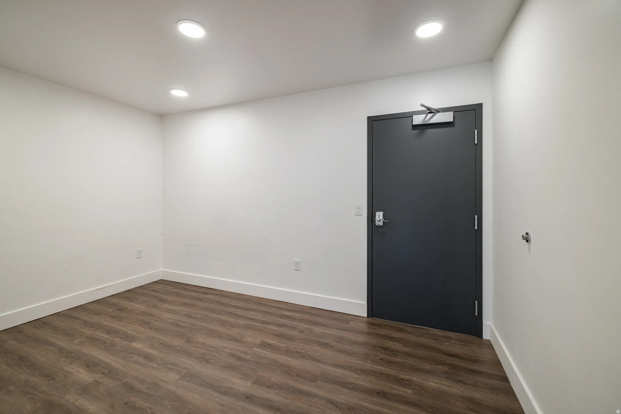 Spacious room featuring wood-finish flooring, recessed lighting, and a dark-toned door with a door closer