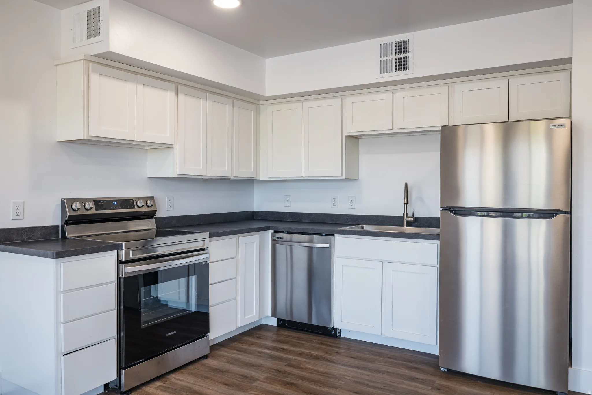 Modern kitchen featuring white shaker cabinetry, dark countertops, stainless steel appliances, and wood-finish flooring