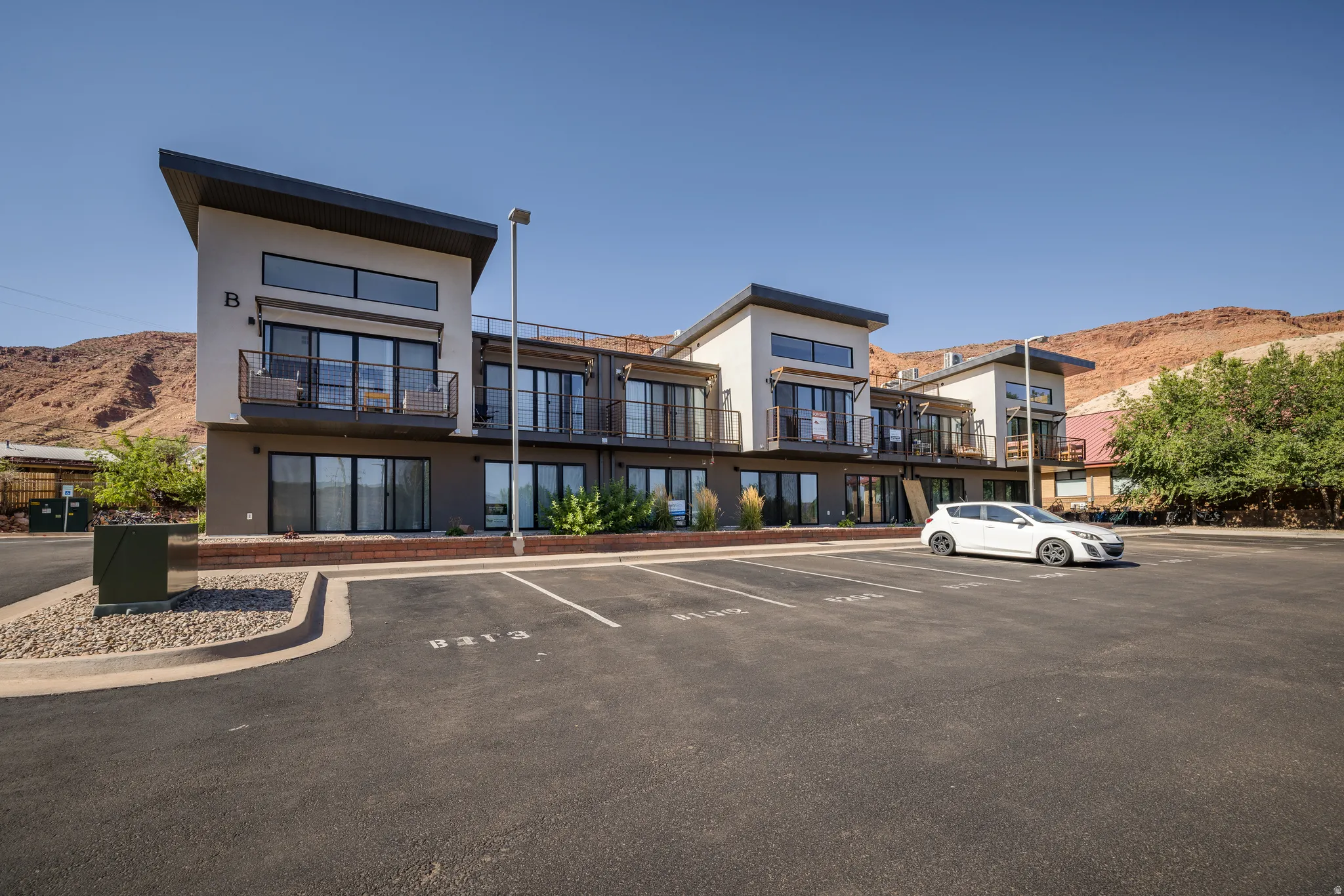 Modern multi-story building featuring light-colored stucco and dark accents, multiple private balconies with black railings, and expansive glass sliding doors