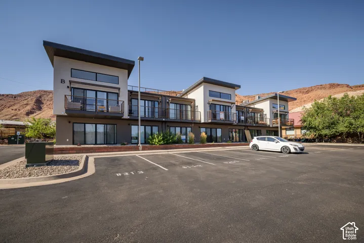 Modern multi-story building featuring light-colored stucco and dark accents, multiple private balconies with black railings, and expansive glass sliding doors