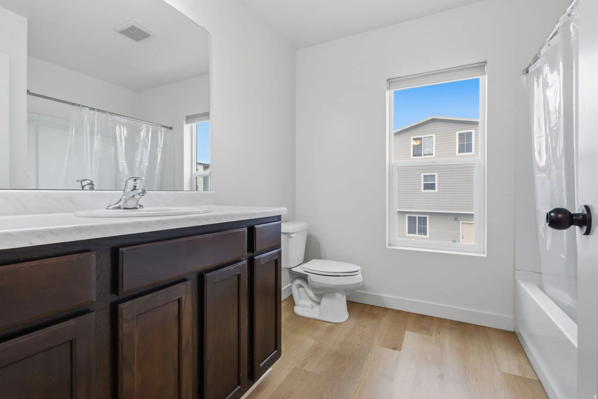 Contemporary bathroom featuring a dark wood-finish vanity with a white countertop and integrated sink, chrome faucet, a toilet, a bathtub with a shower, and light wood-finish flooring