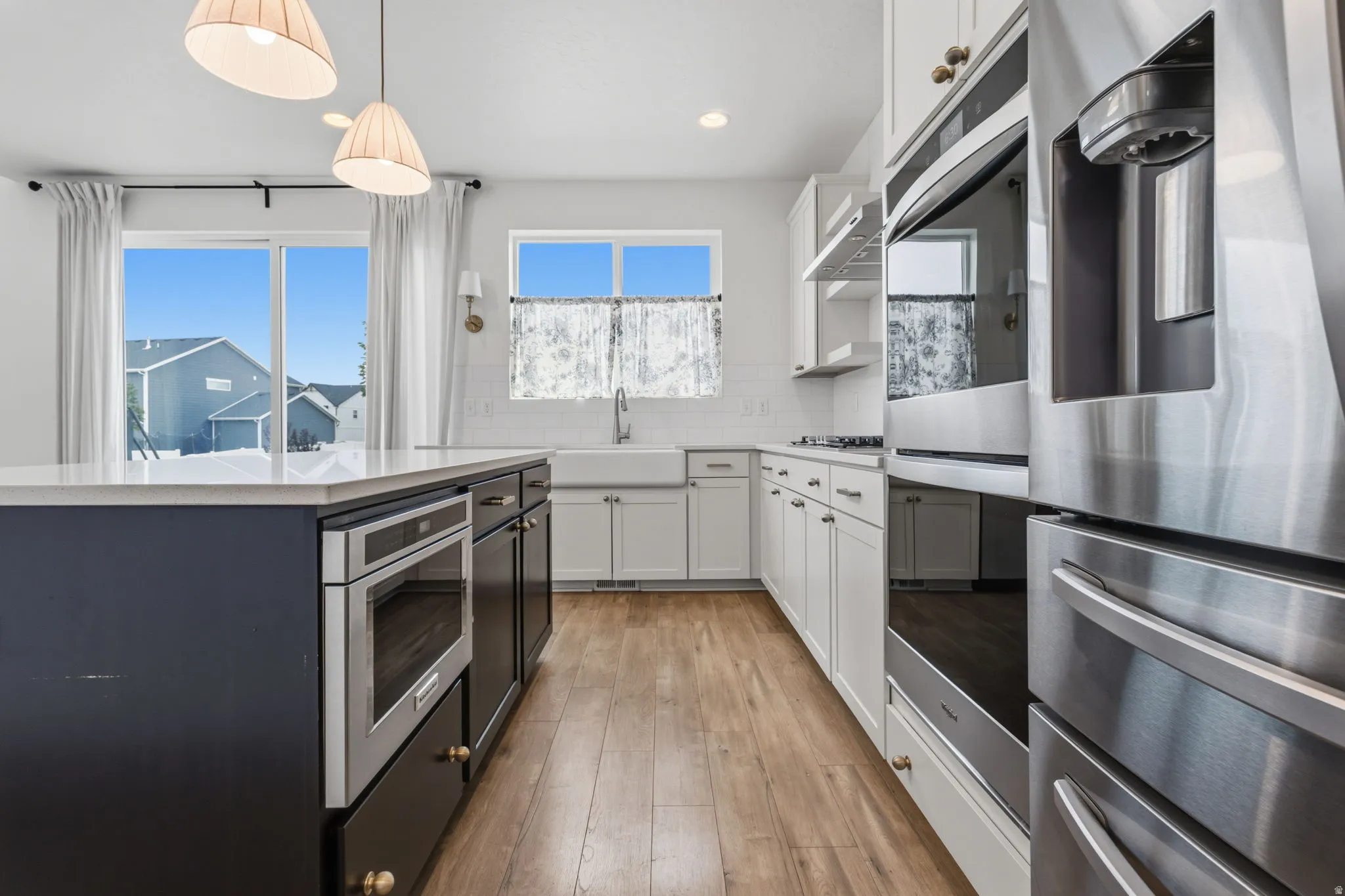 Modern kitchen featuring a large center island with built-in microwave, white shaker cabinetry, stainless steel appliances, wood-finish flooring, and a farmhouse sink