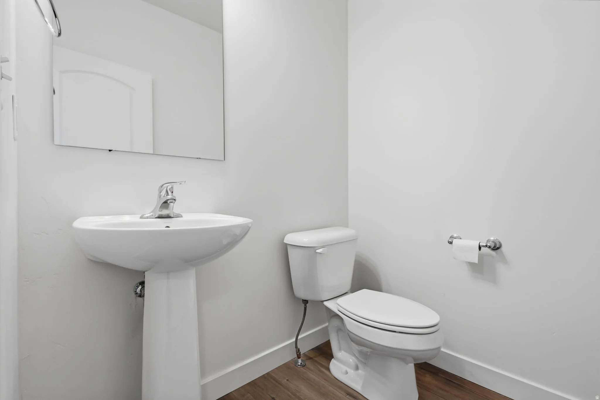 Bathroom featuring a pedestal sink with chrome faucet, a rectangular wall mirror, a white toilet, and wood-finish flooring