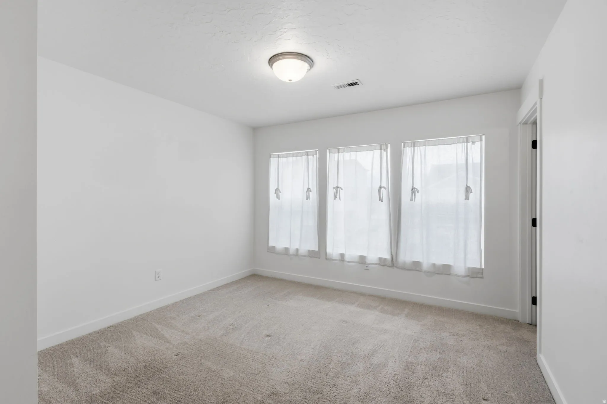 Carpeted room with three windows featuring white curtains, a flush-mount ceiling light fixture, and white wall paint