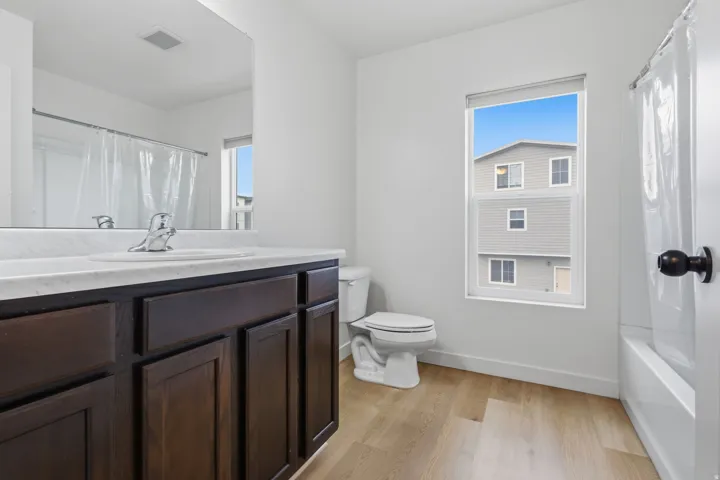 Contemporary bathroom featuring a dark wood-finish vanity with a white countertop and integrated sink, chrome faucet, a toilet, a bathtub with a shower, and light wood-finish flooring