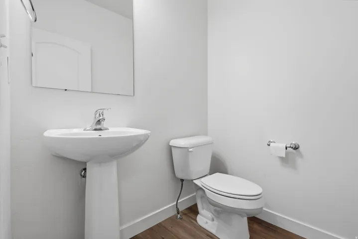 Bathroom featuring a pedestal sink with chrome faucet, a rectangular wall mirror, a white toilet, and wood-finish flooring