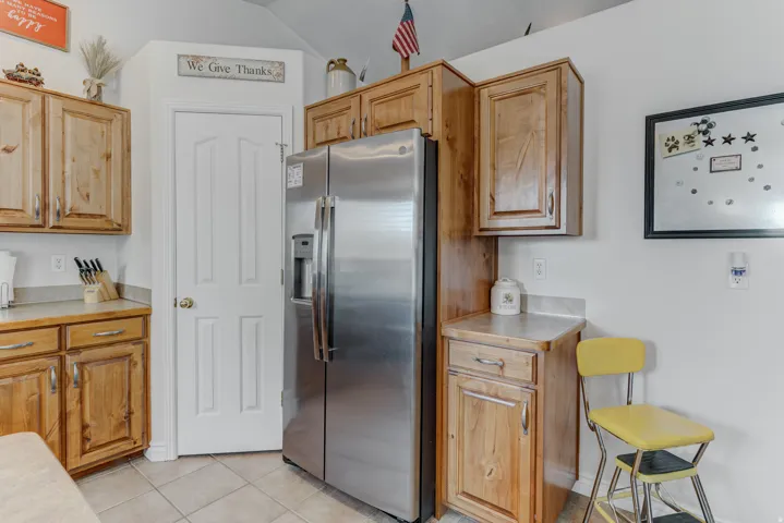 Stainless steel side-by-side refrigerator, light wood-finish cabinetry, light-colored countertops, white paneled door, and neutral tile flooring