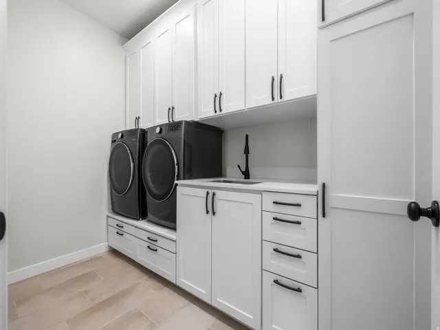Utility room featuring white shaker cabinetry, matte black hardware, a utility sink with a matte black faucet, light-toned tile flooring, and integrated laundry appliances