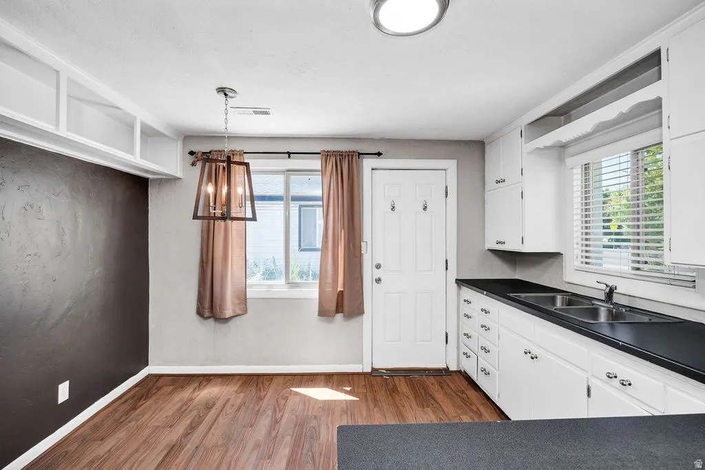 Kitchen area featuring wood-finish flooring, white cabinetry with dark countertops, a double basin sink, and a pendant light fixture