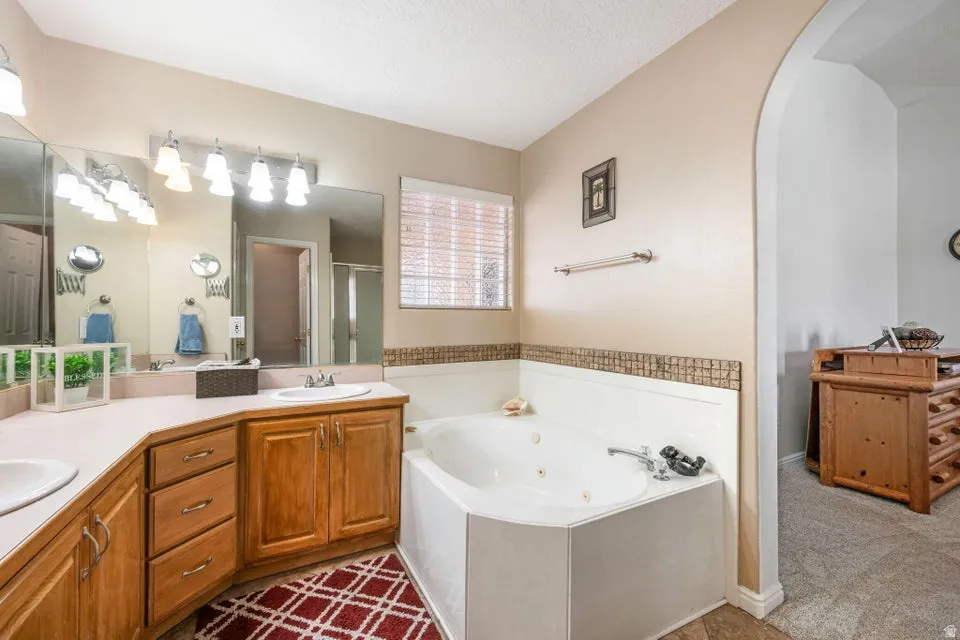 Dual vanity with wood cabinetry, an integrated jetted tub, wall-mounted light fixtures, a window with blinds, and an arched doorway