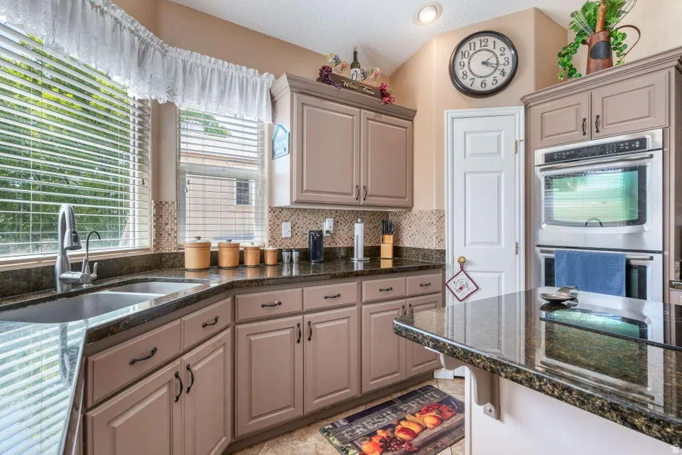 Kitchen featuring recessed lighting, a double basin stainless steel sink with gooseneck faucet, brown cabinetry, dark granite countertops, and a full tile backsplash