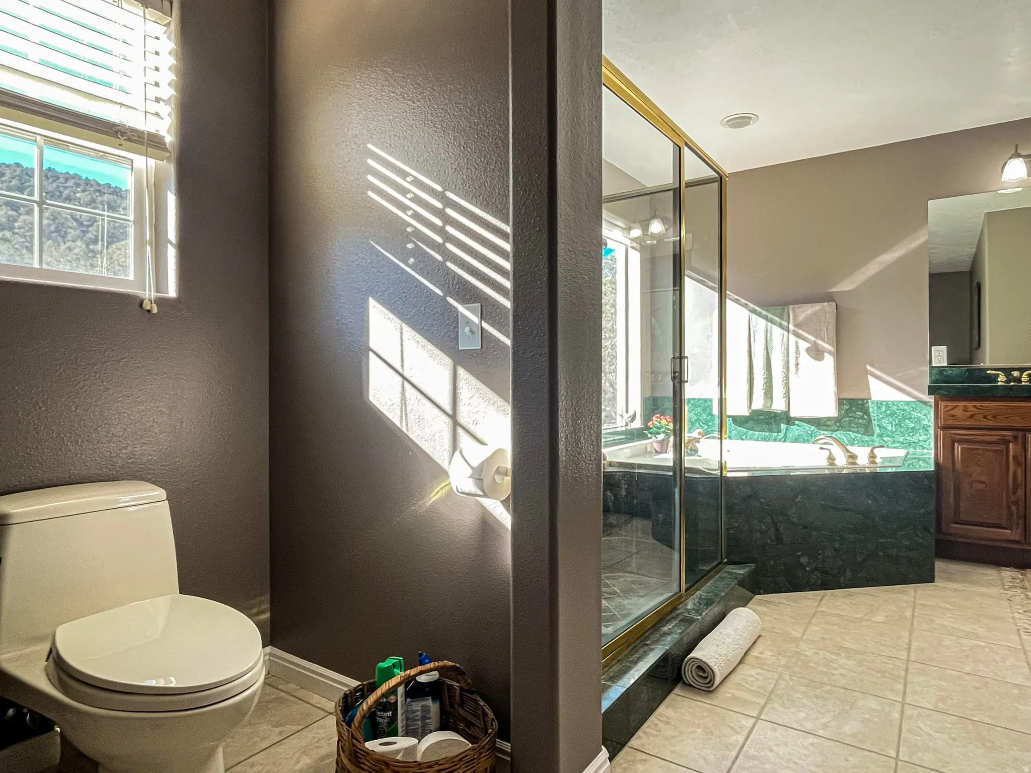 Bathroom featuring a spacious walk-in shower with a gold-framed glass enclosure, deep soaking tub with stone surround, a window with horizontal blinds, and tile flooring
